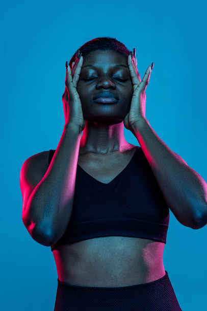 Portrait of young beautiful dark skinned woman posing isolated on blue studio background in neon light. - Photo, image