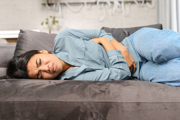 Sad young multiracial woman wearing casual clothes suffering from menstrual pain, feeling sick to her stomach, holding belly, having abdominal cramps during period and lying down - Foto, Imagen