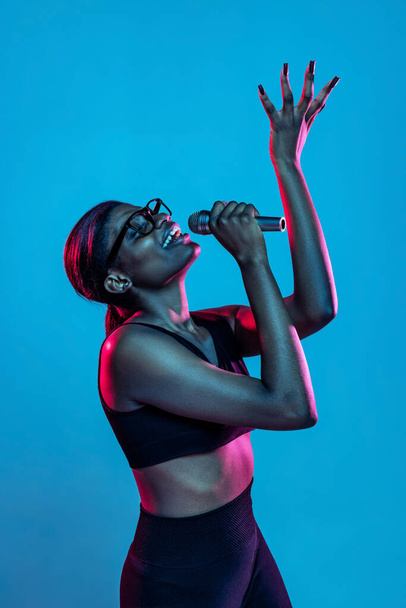 Portrait of young beautiful dark skinned woman posing isolated on blue studio background in neon light. - Фото, зображення