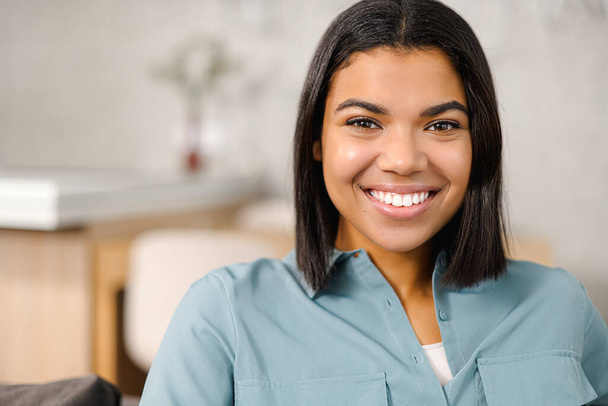Headshot of a young elegant African American ethnic female with beautiful smile looking at the camera while sitting against blurred home interior - Foto, Bild