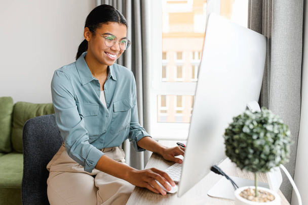 Cheerful multiracial young woman using computer for remote work at home office. Smiling mixed-race businesswoman in casual wear holding video call, typing email, chatting online - Photo, image