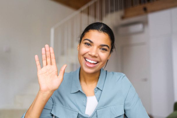 Young and energetic African-American female fresh graduate is ready for an online interview on a video call, sitting at the desk and looking and waving at the camera, saying hello. Job hunting - Φωτογραφία, εικόνα