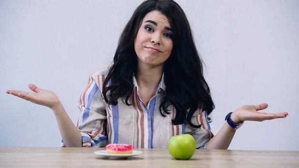 mujer mostrando gesto de encogimiento cerca de manzana y donut aislado en gris - Foto, imagen