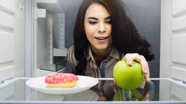 happy woman taking green apple near doughnut in fridge - Foto, Bild