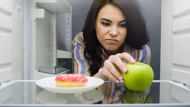 femme mécontente de prendre pomme verte près beignet au réfrigérateur - Photo, image