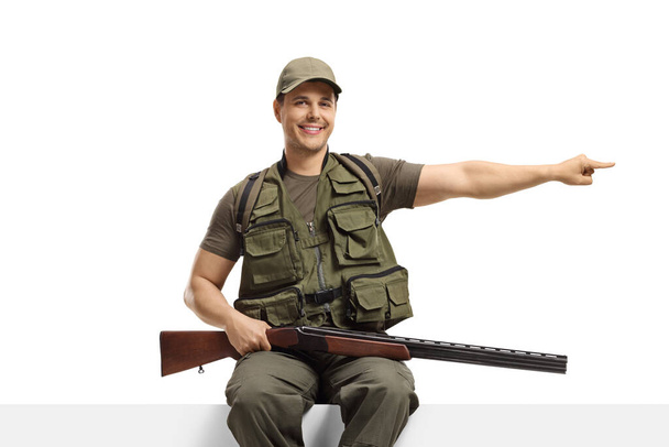 Hunter with a rifle sitting on a panel and pointing to the side isolated on white background - Photo, Image