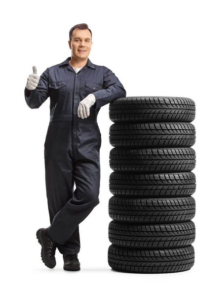 Auto mechanic worker in a uniform leaning on a pile of tires and showing a thumb up gesture isolated on white background - Zdjęcie, obraz
