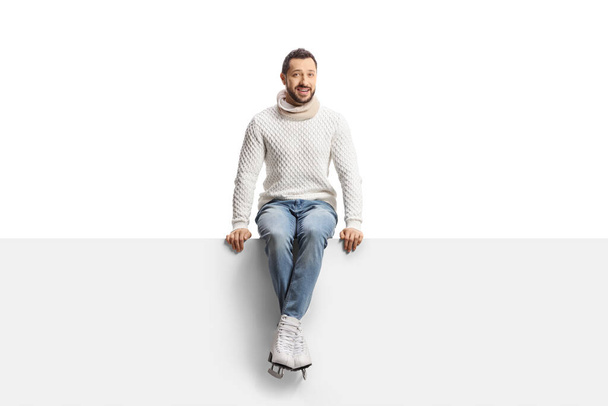 Casual young man wearing ice skates and sitting on a blank panel isolated on white background - Foto, afbeelding