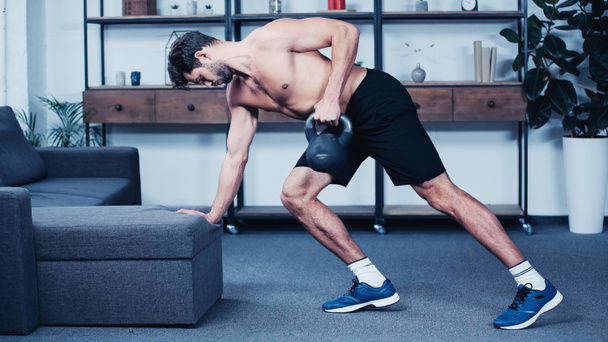 side view of shirtless sportsman training with kettlebell near grey sofa  - Photo, Image