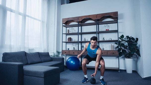 vousatý mladý sportovec v modré tílko cvičení s kettlebell doma - Fotografie, Obrázek