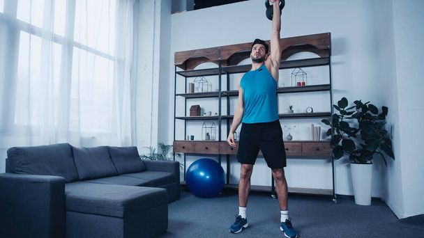 young sportsman in blue tank top exercising with kettlebell at home - Фото, зображення
