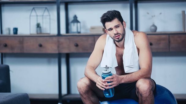 exhausted sportsman with towel sitting on fitness ball and holding sports bottle with water - Foto, immagini