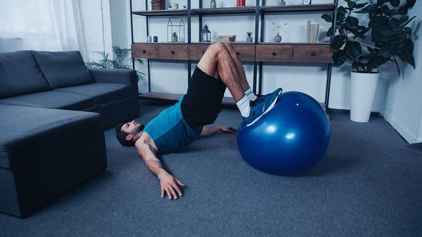 bärtiger Sportler trainiert zu Hause auf blauem Fitnessball  - Foto, Bild