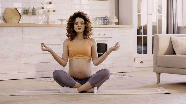 Pregnant woman in sportswear sitting in yoga pose on mat at home  - Foto, Bild