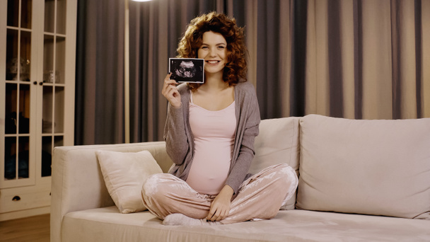 Smiling pregnant woman holding ultrasound scan of baby on couch  - Фото, изображение
