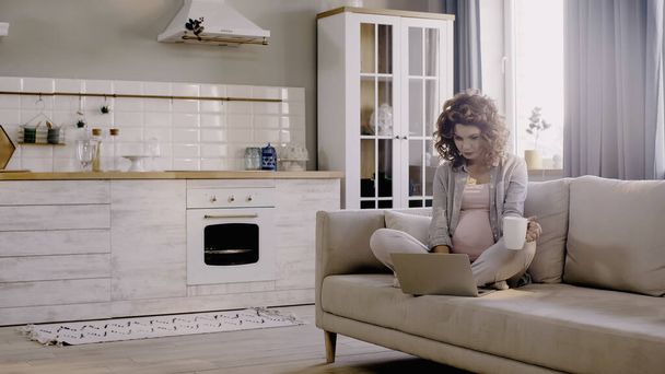 妊娠中のフリーランスはカップを保持し、自宅でソファの上にラップトップを使用  - 写真・画像