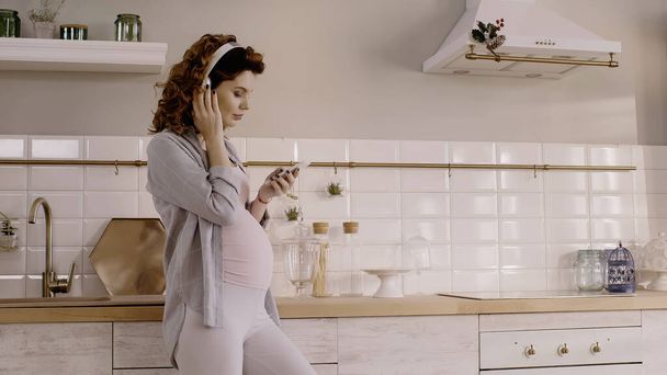 Mujer embarazada joven escuchando música en auriculares y sosteniendo el teléfono inteligente en la cocina  - Foto, Imagen