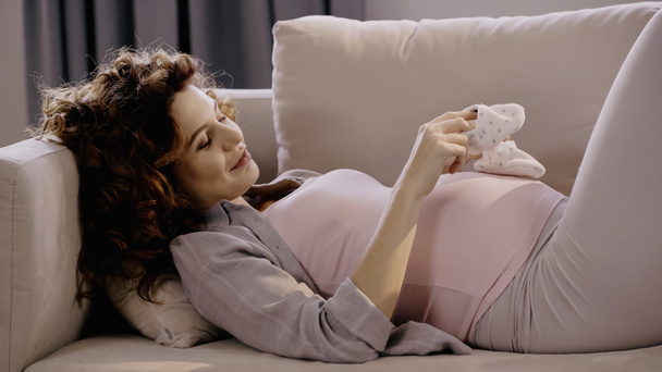 Göndör terhes nő kezében baba csizma közelében has kanapén  - Fotó, kép