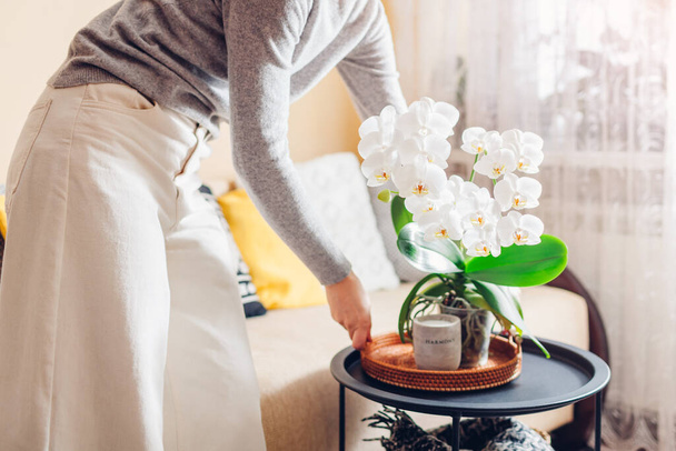 Kobieta kładzie tacę z białymi kwiatami orchidei i świecą na stole dbając o przytulność w domu. Wnętrze i wystrój - Zdjęcie, obraz