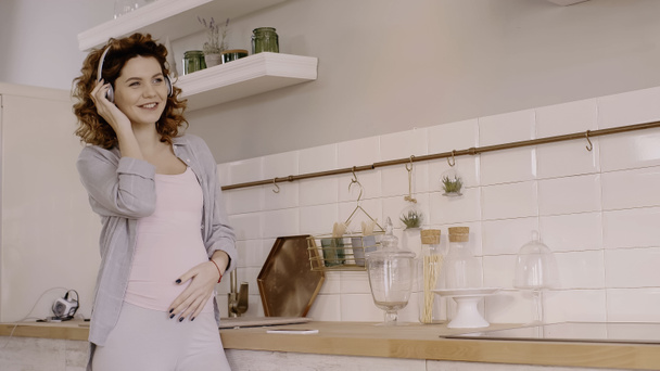 Happy pregnant woman listening music in headphones near cellphone on worktop in kitchen  - Zdjęcie, obraz