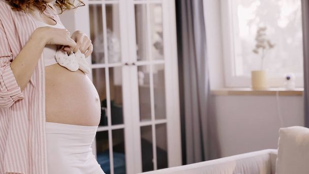Gewassen uitzicht op krullende zwangere vrouw houden baby laarzen in de buurt van buik thuis  - Foto, afbeelding