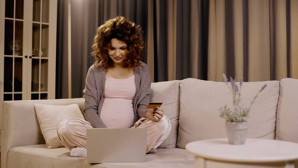 Mujer embarazada con tarjeta de crédito y el uso de la computadora portátil en el sofá  - Foto, Imagen