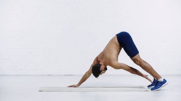 shirtless sportsman stretching back on fitness mat near white brick wall - Foto, afbeelding