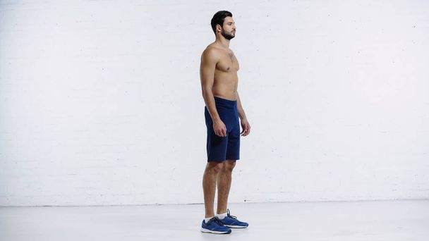 full length of shirtless sportsman in shorts standing near white brick wall  - Zdjęcie, obraz