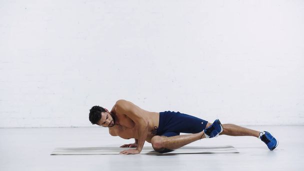 full length of sportsman exercising in one legged plank pose near white brick wall - Photo, Image