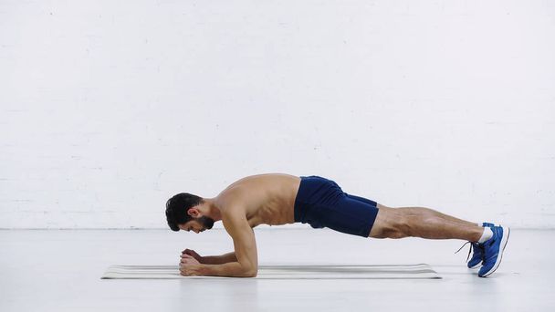 oldalnézetben félmeztelen sportoló csinál deszka gyakorlat fitness szőnyeg közelében fehér tégla fal  - Fotó, kép