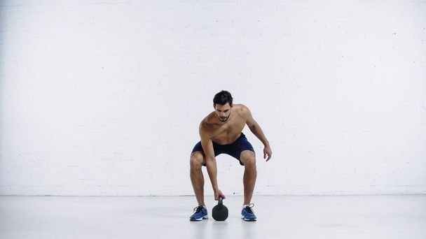 vousatý sportovec cvičení s těžkou kettlebell v blízkosti bílé cihlové zdi  - Fotografie, Obrázek