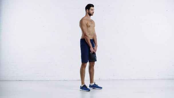 full length of shirtless sportsman in shorts exercising with heavy kettlebell near white brick wall  - Foto, Bild