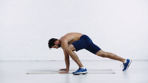 vista lateral del deportista sin camisa haciendo ejercicio en la alfombra de fitness cerca de la pared de ladrillo blanco  - Foto, Imagen