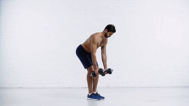 deportista sin camisa en pantalones cortos de entrenamiento con pesadas mancuernas cerca de la pared de ladrillo blanco - Foto, imagen