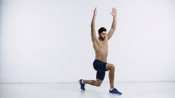 full length of shirtless sportsman doing lunges with raised hands near white brick wall  - Photo, Image