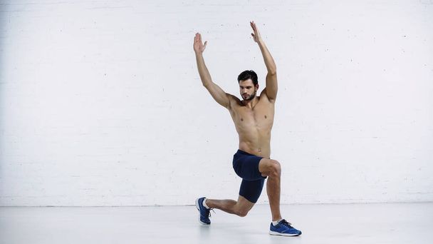 full length of bearded young sportsman doing lunges with raised hands near white brick wall  - Photo, Image