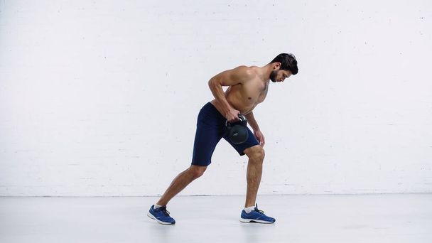 sportsman in sneakers exercising with heavy kettlebell near white brick wall  - Photo, Image