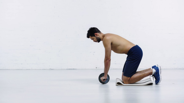 side view of shirtless sportsman exercising with abdominal wheel near white brick wall - Foto, imagen