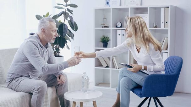 blonde psychologist giving glass of water to middle aged man in sportswear in consulting room  - Foto, immagini