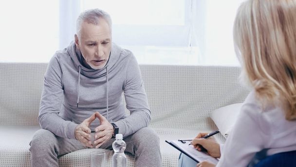 triste hombre de mediana edad en ropa deportiva sentado en el sofá cerca de psicoterapeuta borrosa en la clínica  - Foto, Imagen