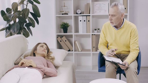 blonde woman lying on couch and looking at middle aged psychotherapist with clipboard  - Photo, Image
