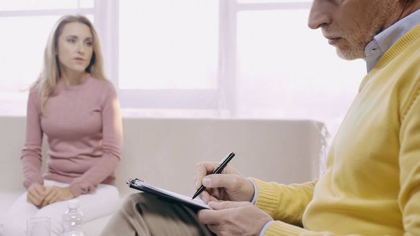 psychotherapeut van middelbare leeftijd die schrijven op klembord en luisteren wazig patiënt zitten op de bank in consulting room  - Foto, afbeelding