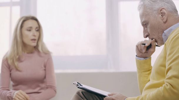 pensativo psicoterapeuta de mediana edad sosteniendo pluma cerca del portapapeles y escuchando paciente borroso en la sala de consulta  - Foto, imagen