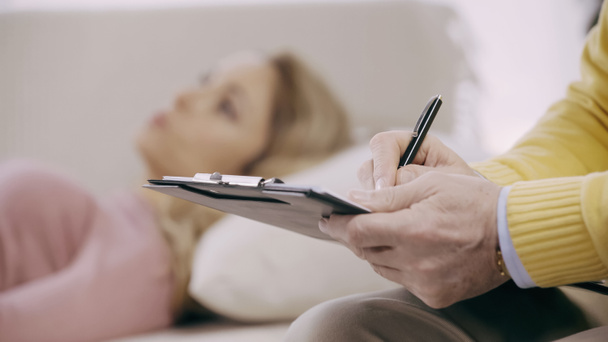 close up of mature psychotherapist writing on clipboard near blurred patient  - Photo, Image