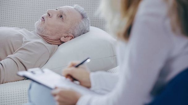 middle aged man lying on couch and talking during consultation with blurred psychologist holding clipboard - Fotografie, Obrázek