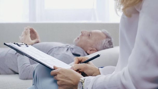 psychologist holding clipboard near blurred middle aged man lying on sofa during consultation  - Valokuva, kuva