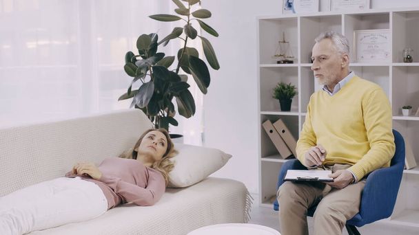blonde woman lying on couch near pensive middle aged psychotherapist with clipboard - Photo, Image
