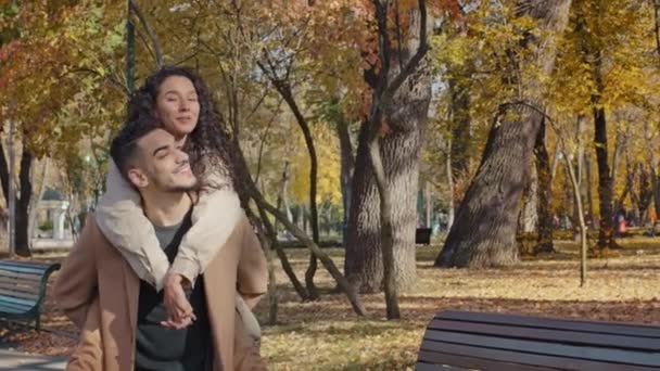 Young guy carries girl on back playful hispanic couple walking in autumn park girl laughing keep stretched out arms at side play airplanes fun romantic date outdoors family dream of traveling together - Filmagem, Vídeo