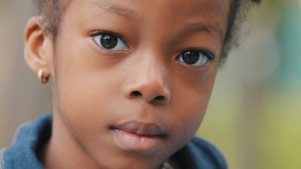 Lähikuva mietteliäs pieni tyttö afrikkalainen amerikkalainen lapsi nostaa päänsä katsoen kameran edessä näkymä surullinen vauva ulkona muotokuva hymyilemätön koulutyttö kaunis tunteeton kasvot yksinäinen söpö lapsi tylsistynyt ilme - Materiaali, video