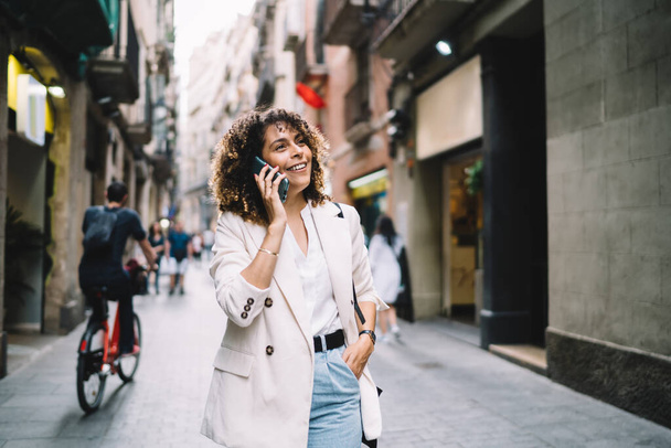 Pretty female tourist in stylish clothing walking at city street and discussing solo vacations experience during international smartphone conversation in roaming, happy girl using cell for talking - Φωτογραφία, εικόνα